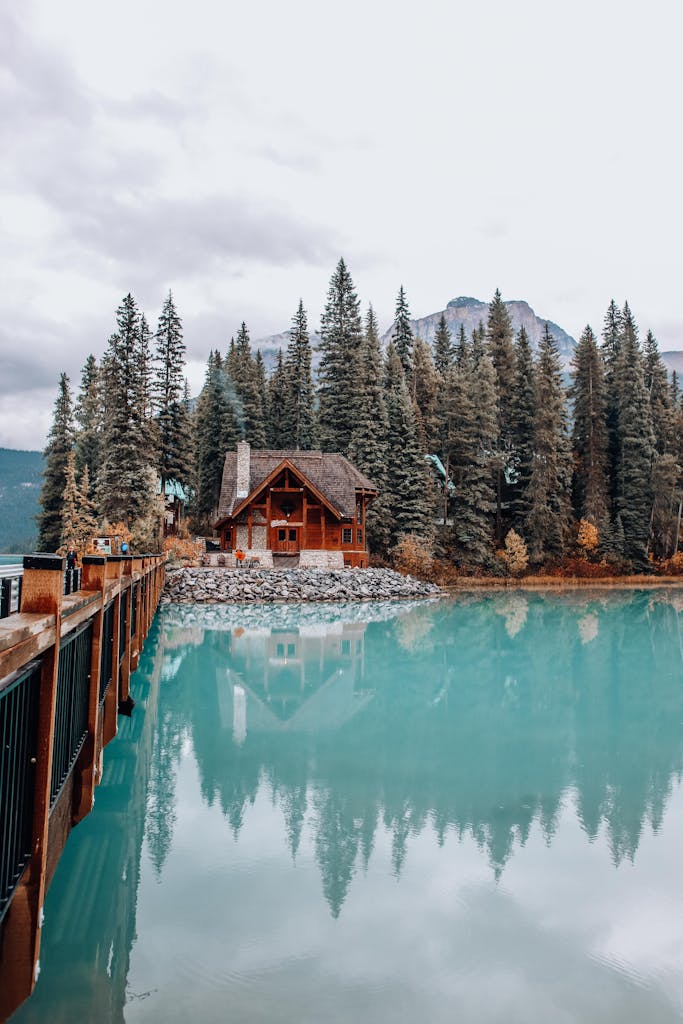 Charming wooden house reflected in calm lake, surrounded by lush forest and mountains.